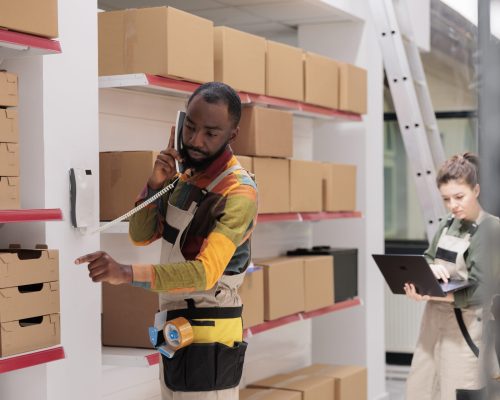 African american manager discussing merchandise logistics with remote supervisor at landline phone in warehouse. Employee working at customers orders, checking shipment details in storehouse
