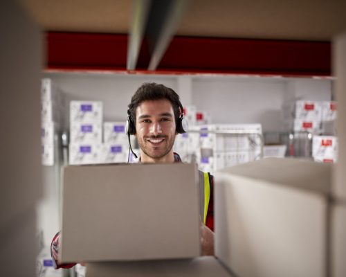 Portrait Of Male Worker Wearing Headset In Logistics Distribution Warehouse Taking Box From Shelf